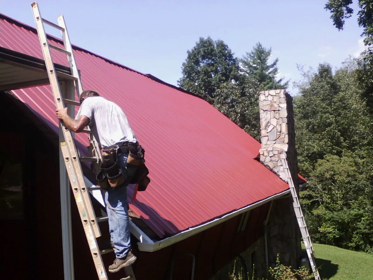 Roofer on a ladder beside a red metal roof and stone chimney — Soffit & Fascia Repair by Roofing Brothers in Vinton